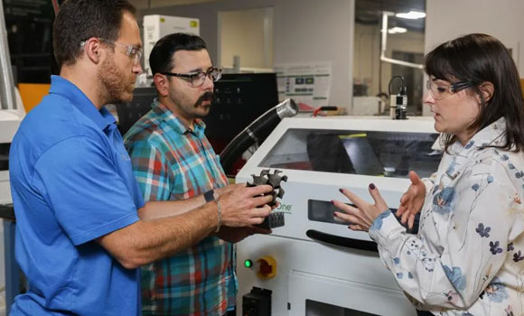 From left, Corson Cramer, Trevor Aguirre and Amy Elliott discuss the silicon carbide gyroid component, additively manufactured using the Binder Jetting machine seen in the background (Courtesy ORNL)